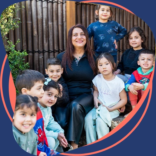 Teacher sitting with a group of smiling children outdoors, representing a fun learning environment.